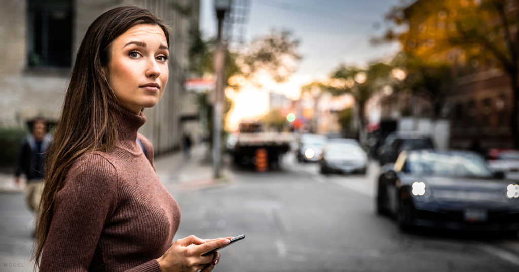 A woman who had breast augmentation surgery stands on the sidewalk holding her cell phone.